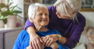 A middle-aged woman standing behind her senior mother with her arms around her in a comforting hug, smiling at each other