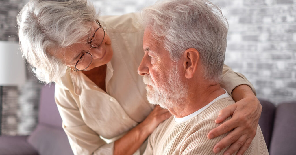 A senior woman standing beside a seated senior man with her hands on his shoulders, comforting him