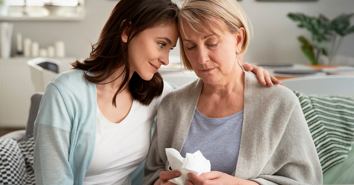 Two women sitting together on a sofa, one comforting the other as she experiences grief.